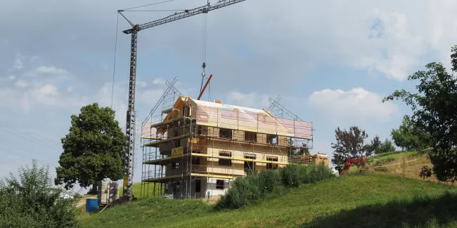 Ein mehrstöckiges Holzhaus im Bau, umgeben von Gerüsten und einem Baukran, steht auf einer grünen Anhöhe unter blauem Himmel mit einigen Wolken.