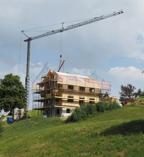 Ein mehrstöckiges Holzhaus im Bau, umgeben von Gerüsten und einem Baukran, steht auf einer grünen Anhöhe unter blauem Himmel mit einigen Wolken.