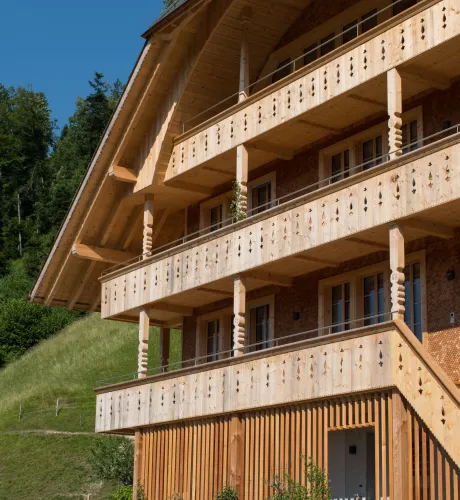 Mehrstöckiges Holzhaus im traditionellen Schweizer Stil mit kunstvoll verzierten Holzbalkonen und grosszügigem Dachüberstand. Die Fassade besteht aus hellem, massivem Holz, umgeben von einer grünen Hanglandschaft bei klarem Himmel.