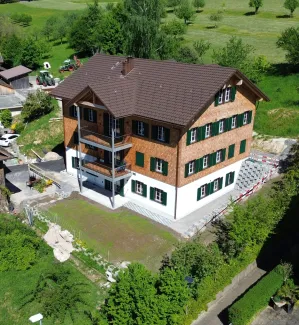 Luftaufnahme eines frisch renovierten, mehrstöckigen Holzhauses im traditionellen Stil mit brauner Schindelfassade und grünen Fensterläden. Das Gebäude steht inmitten einer ländlichen Umgebung mit Wiesen, Bäumen und benachbarten Wohn- und Wirtschaftsbauten. Vor dem Haus befindet sich ein neu angelegter Vorplatz, während rundherum noch Bereiche der Gartengestaltung und Bauarbeiten sichtbar sind. Im Hintergrund erstreckt sich eine grüne Hügellandschaft mit verstreuten Obstbäumen und einigen modernen Wohnhäuse