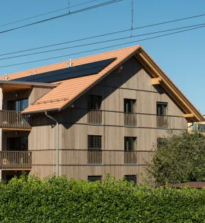 Aussenansicht eines mehrgeschossigen Wohnhauses in Holzbauweise mit Satteldach und integrierten Solarpanels. Die Fassade besteht aus senkrechter Holzschalung, die Balkone sind aus Holz gefertigt. Das Gebäude steht in einem Wohnquartier mit Hecken und Nachbarbauten, im Vordergrund verlaufen Bahnoberleitungen vor blauem Himmel.