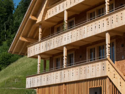 Mehrstöckiges Holzhaus im traditionellen Schweizer Stil mit kunstvoll verzierten Holzbalkonen und grosszügigem Dachüberstand. Die Fassade besteht aus hellem, massivem Holz, umgeben von einer grünen Hanglandschaft bei klarem Himmel.