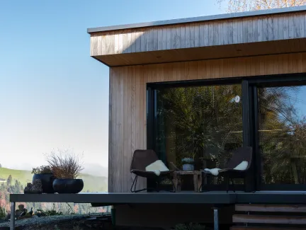 Aussenansicht eines modernen Holzhauses mit klarer Linienführung. Auf der erhöhten Terrasse stehen zwei Stühle mit Kissen und ein kleiner Tisch vor einer grossen Glasfront. Die Fassade besteht aus senkrechter Holzschalung, im Hintergrund sind Hügel, Bäume und eine ländliche Landschaft bei klarem Himmel zu sehen.