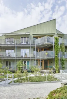 Aussenansicht eines modernen, mehrstöckigen Wohngebäudes in Holzbauweise mit hellgrüner Fassade. Das Haus verfügt über grosse Balkone mit Metallgeländern und Kletterpflanzen, die vertikal an den Fassadenelementen emporwachsen. Vor dem Gebäude liegt ein gepflegter Garten mit jungen Bäumen, Sträuchern und geschwungenen Wegen. Die Architektur wirkt offen und einladend, mit vielen Fenstern und einer klar strukturierten Formensprache. Im Vordergrund sind ein Kiesweg und ein Zaun sichtbar, während der Himmel leic