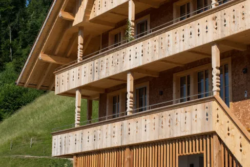 Mehrstöckiges Holzhaus im traditionellen Schweizer Stil mit kunstvoll verzierten Holzbalkonen und grosszügigem Dachüberstand. Die Fassade besteht aus hellem, massivem Holz, umgeben von einer grünen Hanglandschaft bei klarem Himmel.