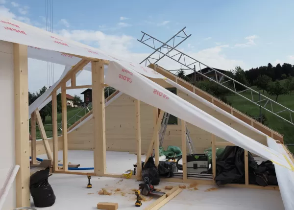 Eine Holzrahmenkonstruktion eines Dachgeschosses im Bau, teilweise mit Dampfsperrfolie abgedeckt, umgeben von Werkzeugen und Baumaterial auf einer Baustelle im Grünen.
