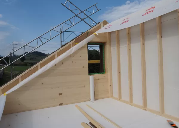Innenansicht eines Holzrahmenbaus mit schräger Dachwand, sichtbarer Holzverkleidung und Dämmfolie, im Hintergrund grüne Hügel und Gerüst im Freien.