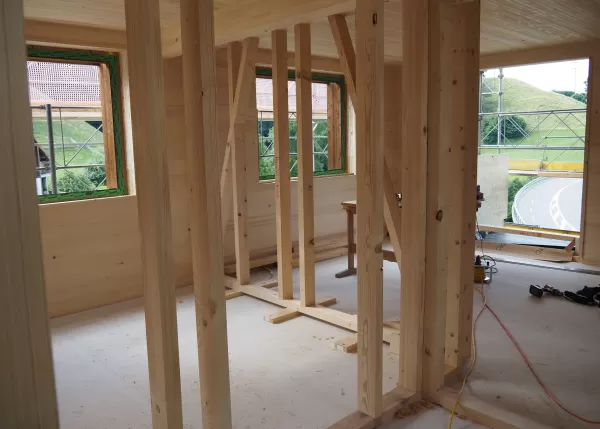 Innenansicht eines Holzhauses im Rohbau mit sichtbaren Wand- und Deckenbalken, Fensterausschnitten und Blick auf das umliegende Gerüst und die Straße.