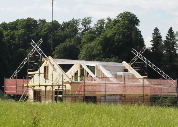 Neubau eines Holzhauses mit Gerüst und offener Dachkonstruktion auf einer grünen Wiese, umgeben von Bäumen und blauem Himmel.