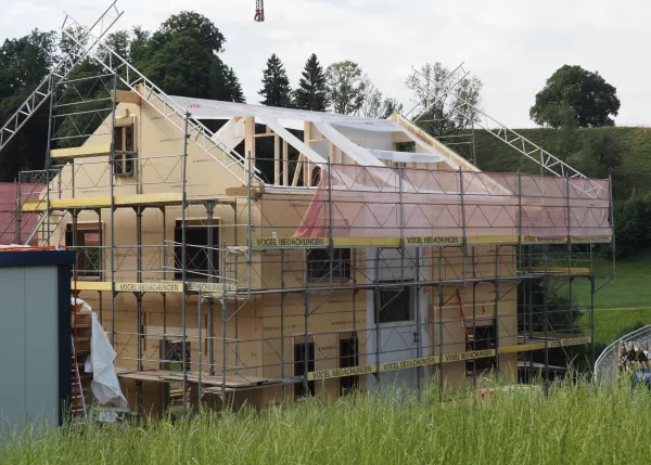 Holzhaus im Rohbau mit Gerüst und Dachaufbau, umgeben von grüner Landschaft und sanften Hügeln.