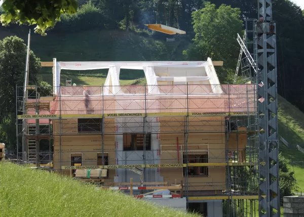 Ein mehrstöckiges Holzhaus im Bau, umgeben von Gerüsten und einem Baukran, steht auf einer grünen Anhöhe unter blauem Himmel mit einigen Wolken.