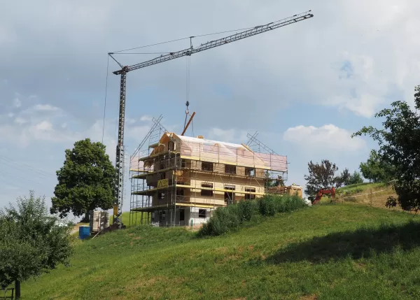 Ein mehrstöckiges Holzhaus im Bau, umgeben von Gerüsten und einem Baukran, steht auf einer grünen Anhöhe unter blauem Himmel mit einigen Wolken.