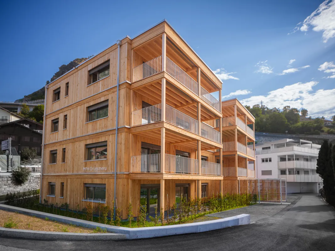 Aussenansicht eines modernen, viergeschossigen Mehrfamilienhauses in Holzbauweise vor einer felsigen Bergkulisse. Das Gebäude verfügt über eine vertikale Holzfassade sowie grosszügige, überdachte Balkone mit sichtbarer Holzkonstruktion und Metallgeländern.