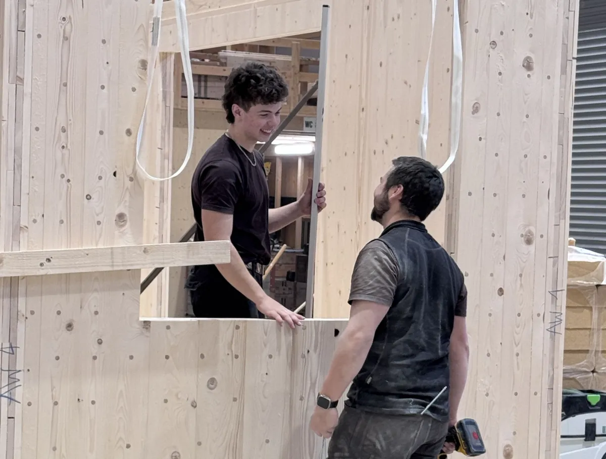 Zwei Mitarbeitende der Zimmerei Hirschi montieren in der Werkhalle ein vorgefertigtes Holzelement. Sie arbeiten an einer massiven Holzrahmenkonstruktion mit Fensteröffnung und stimmen sich dabei direkt vor Ort ab.