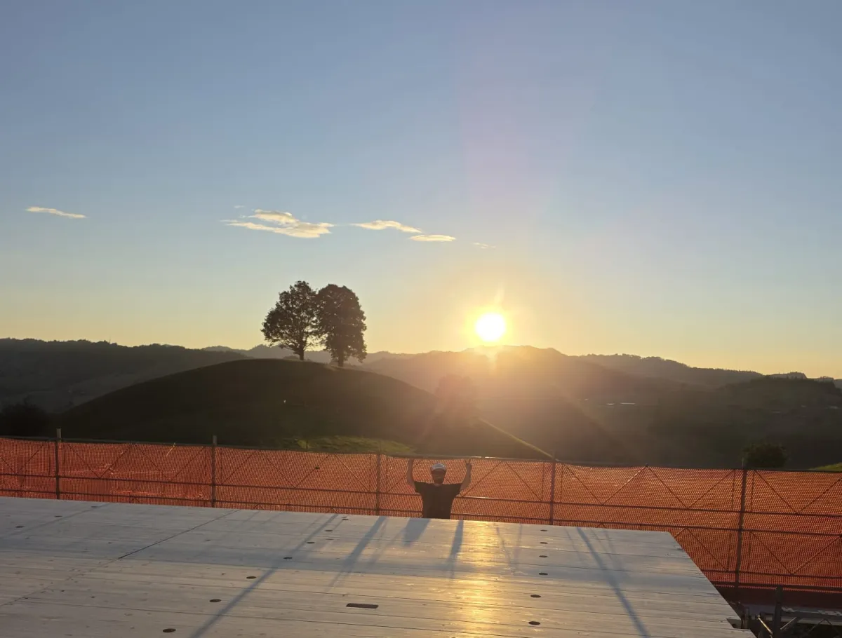 Blick über eine neu verlegte Holzdecke auf einer Baustelle bei Sonnenuntergang. Im Hintergrund steht ein Zimmermann am Gerüst vor der hügeligen Landschaft und hebt beide Arme in die Höhe, während die tief stehende Sonne über den Hügeln scheint.