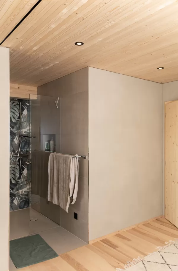 Truberholz Einfamilienhaus mit integrierter Arven-Täfer Wand. Schlafzimmer mit integrierter Dusche und Waschtisch. In der Dusche ist ein Brettstappel-Decke und der Boden ist aus ein Eschen Riemenboden. Die Wände sind Truberholz sichtbar. Die Decke ist leicht in der Dachschräge und ist eine Massivholzplatte. In der Wohnstube ist ein Cheminée mit Sitzoffen/Sitzbank.