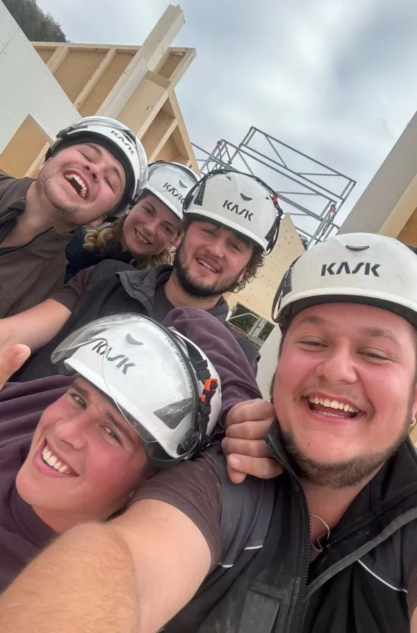 Baustelle eines Holzbaus bei Sonnenuntergang. Die beleuchtete Holzkonstruktion und das eingerüstete Gebäude zeigen den fortgeschrittenen Aufbau eines modernen Holzhauses durch die Zimmerei Hirschi AG.