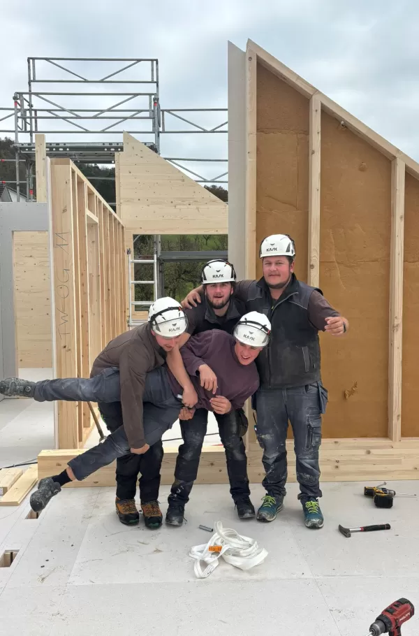 Baustelle eines Holzbaus bei Sonnenuntergang. Die beleuchtete Holzkonstruktion und das eingerüstete Gebäude zeigen den fortgeschrittenen Aufbau eines modernen Holzhauses durch die Zimmerei Hirschi AG.