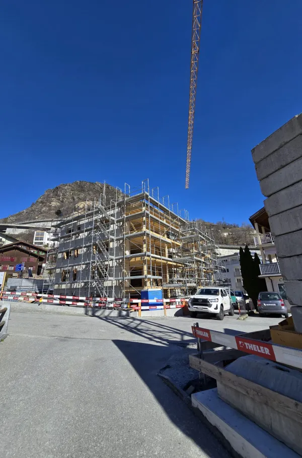 Zimmerleute arbeiten an der Holzrahmenkonstruktion eines mehrgeschossigen Gebäudes auf einer Baustelle. Die tragenden Wände aus Holz werden vor Ort montiert, während ein Baukran über dem Bauplatz steht. Im Hintergrund erstrecken sich Dorfhäuser und eine beeindruckende alpine Berglandschaft