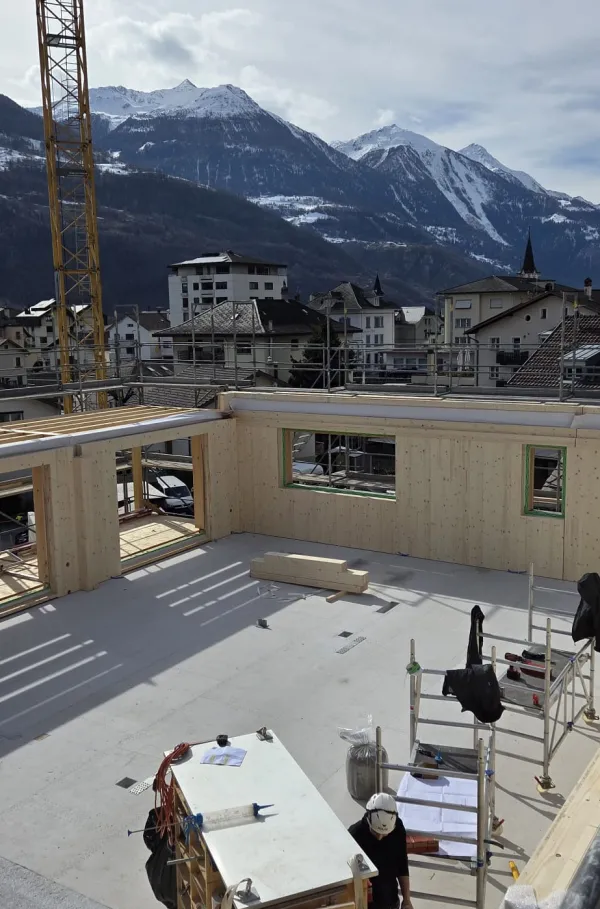 Zimmerleute arbeiten an der Holzrahmenkonstruktion eines mehrgeschossigen Gebäudes auf einer Baustelle. Die tragenden Wände aus Holz werden vor Ort montiert, während ein Baukran über dem Bauplatz steht. Im Hintergrund erstrecken sich Dorfhäuser und eine beeindruckende alpine Berglandschaft