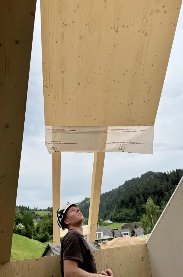 Zimmerleute montieren während der Aufrichtearbeiten eine vorgefertigte Holzkonstruktion auf einem Gebäude. Ein Kran hebt ein grosses Dachelement an seinen Platz, während mehrere Fachkräfte auf dem Dach die Montage begleiten.