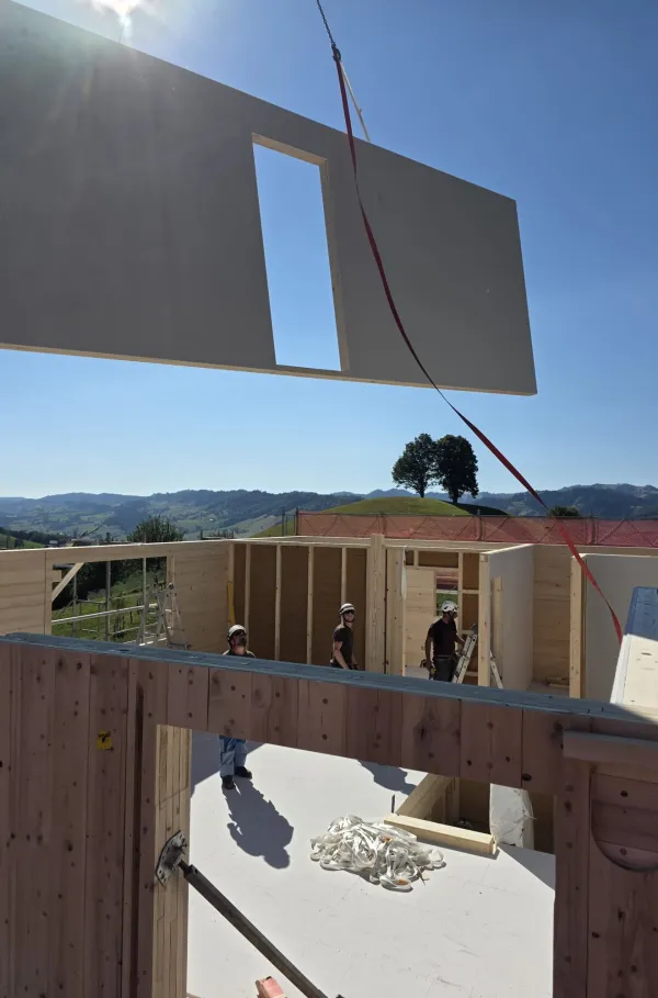 Blick über eine neu verlegte Holzdecke auf einer Baustelle bei Sonnenuntergang. Im Hintergrund steht ein Zimmermann am Gerüst vor der hügeligen Landschaft und hebt beide Arme in die Höhe, während die tief stehende Sonne über den Hügeln scheint