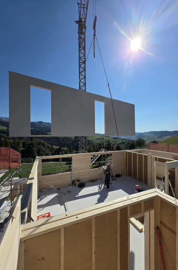 Blick über eine neu verlegte Holzdecke auf einer Baustelle bei Sonnenuntergang. Im Hintergrund steht ein Zimmermann am Gerüst vor der hügeligen Landschaft und hebt beide Arme in die Höhe, während die tief stehende Sonne über den Hügeln scheint