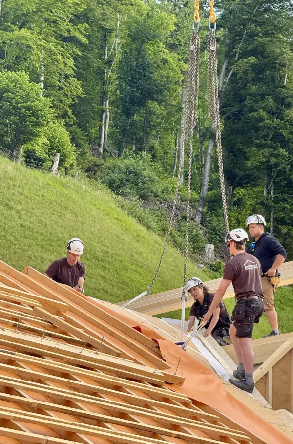 Zimmerleute montieren während der Aufrichtearbeiten eine vorgefertigte Holzkonstruktion auf einem Gebäude. Ein Kran hebt ein grosses Dachelement an seinen Platz, während mehrere Fachkräfte auf dem Dach die Montage begleiten.