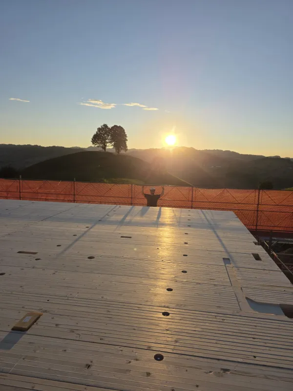 Blick über eine neu verlegte Holzdecke auf einer Baustelle bei Sonnenuntergang. Im Hintergrund steht ein Zimmermann am Gerüst vor der hügeligen Landschaft und hebt beide Arme in die Höhe, während die tief stehende Sonne über den Hügeln scheint
