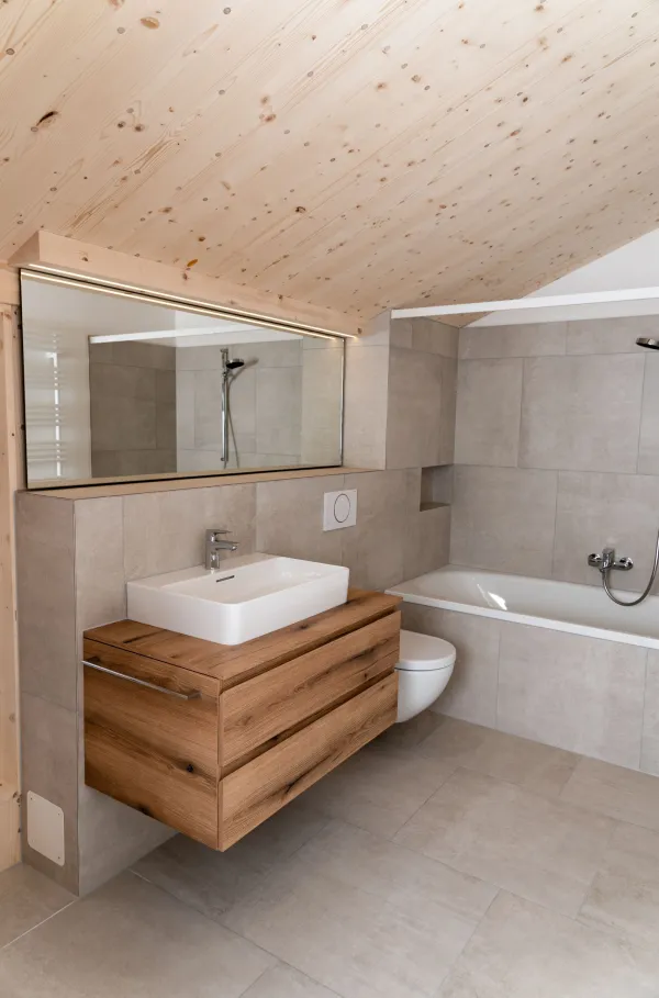 Modernes Badezimmer mit grossformatigen grauen Fliesen, Badewanne und Waschtisch mit hölzernem Unterschrank. Die helle, geneigte Holzdecke und die sichtbaren Holzelemente schaffen einen warmen Kontrast zur schlichten Badezimmereinrichtung.