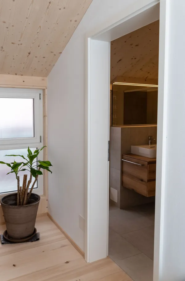 Modernes Badezimmer mit grossformatigen grauen Fliesen, Badewanne und Waschtisch mit hölzernem Unterschrank. Die helle, geneigte Holzdecke und die sichtbaren Holzelemente schaffen einen warmen Kontrast zur schlichten Badezimmereinrichtung.