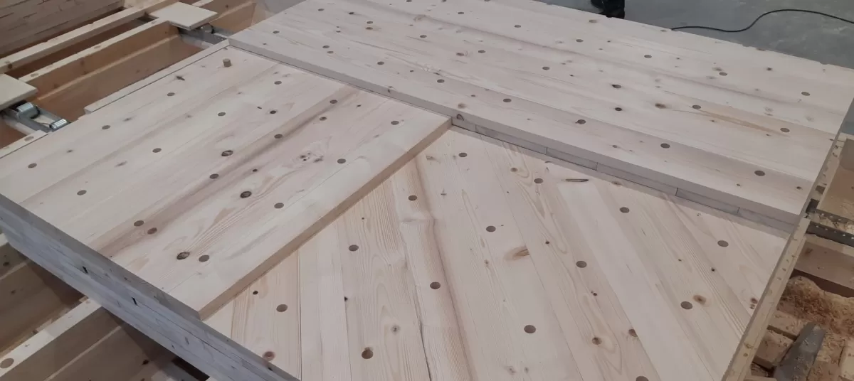 In unserer Produktionsstätte in Trub haben die Arbeiten zu den Wandelementen begonnen. Die Fertigungsanlage umfasst einen Produktionstisch, ein Dübelportal und ein Abbundportal. Die auf dem Produktionstisch ausgelegten Bretter werden vom Dübelportal vollautomatisch bei den Kreuzungspunkten verbunden. Das Abbundportal fräst die Konturen, bohrt die Steckdosen, kehlt die Verbindungsnuten und kalibriert die Elemente auf die gewünschte Dicke.