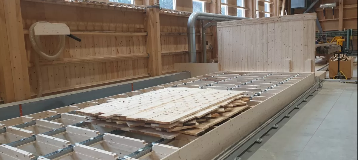 In unserer Produktionsstätte in Trub haben die Arbeiten zu den Wandelementen begonnen. Die Fertigungsanlage umfasst einen Produktionstisch, ein Dübelportal und ein Abbundportal. Die auf dem Produktionstisch ausgelegten Bretter werden vom Dübelportal vollautomatisch bei den Kreuzungspunkten verbunden. Das Abbundportal fräst die Konturen, bohrt die Steckdosen, kehlt die Verbindungsnuten und kalibriert die Elemente auf die gewünschte Dicke.