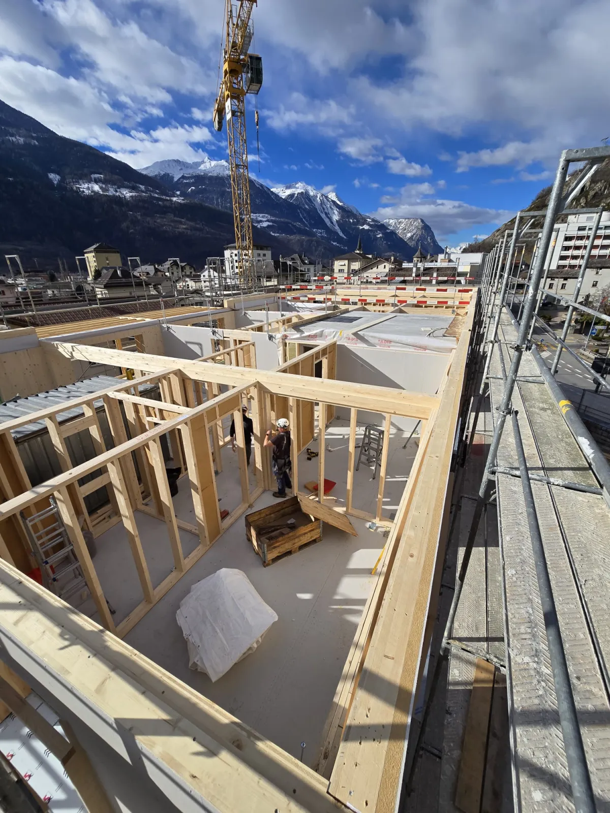 Zimmerleute arbeiten an der Holzrahmenkonstruktion eines mehrgeschossigen Gebäudes auf einer Baustelle. Die tragenden Wände aus Holz werden vor Ort montiert, während ein Baukran über dem Bauplatz steht. Im Hintergrund erstrecken sich Dorfhäuser und eine beeindruckende alpine Berglandschaft.