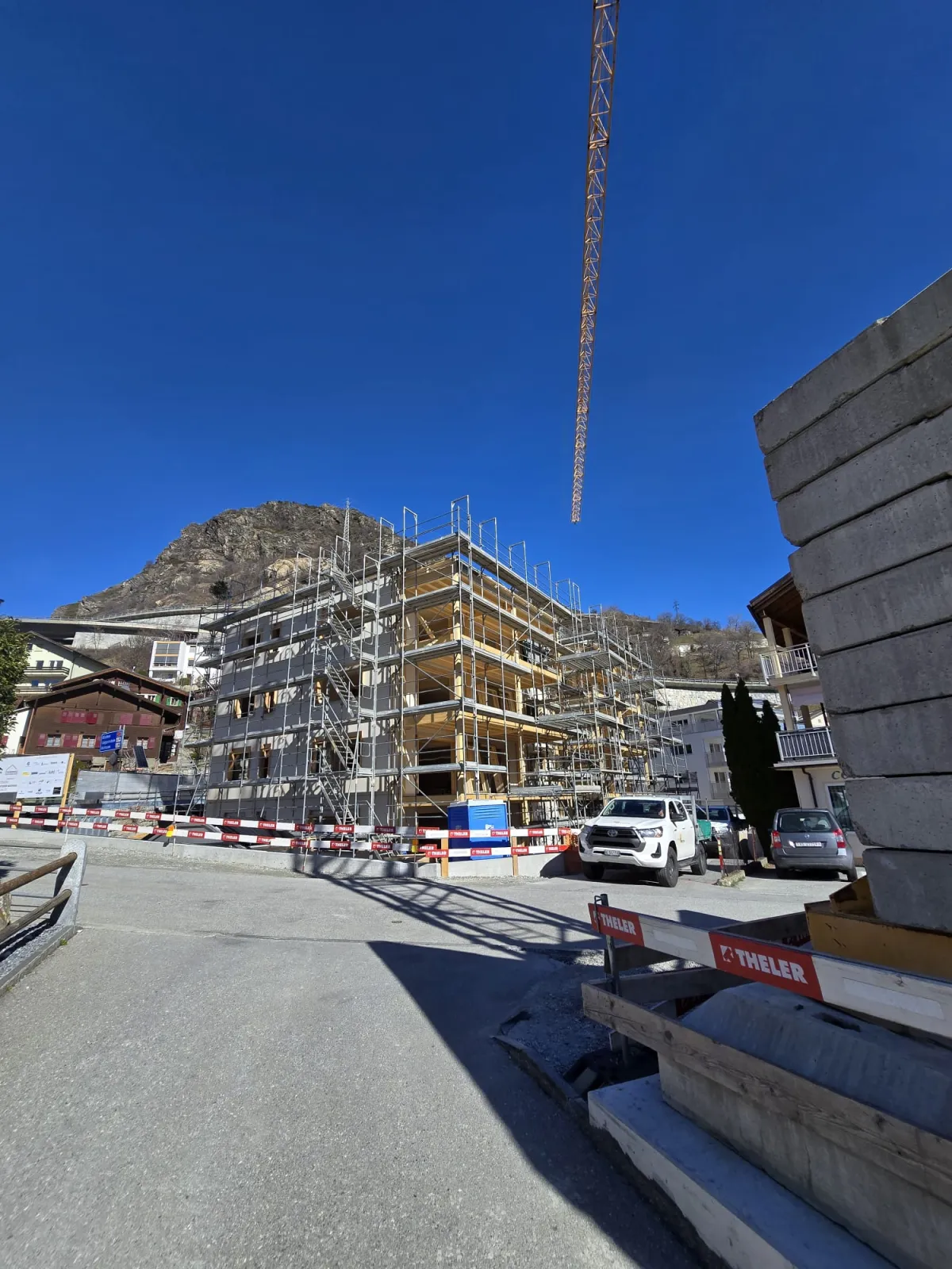 Zimmerleute arbeiten an der Holzrahmenkonstruktion eines mehrgeschossigen Gebäudes auf einer Baustelle. Die tragenden Wände aus Holz werden vor Ort montiert, während ein Baukran über dem Bauplatz steht. Im Hintergrund erstrecken sich Dorfhäuser und eine beeindruckende alpine Berglandschaft.