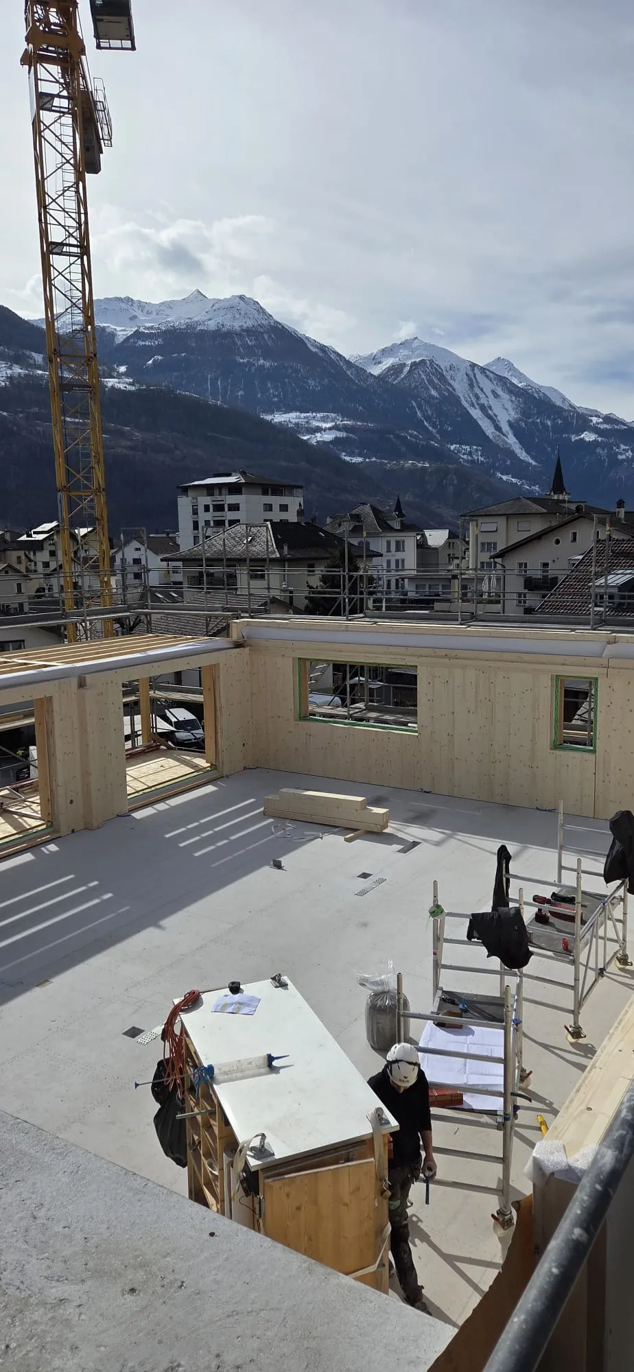 Zimmerleute arbeiten an der Holzrahmenkonstruktion eines mehrgeschossigen Gebäudes auf einer Baustelle. Die tragenden Wände aus Holz werden vor Ort montiert, während ein Baukran über dem Bauplatz steht. Im Hintergrund erstrecken sich Dorfhäuser und eine beeindruckende alpine Berglandschaft.