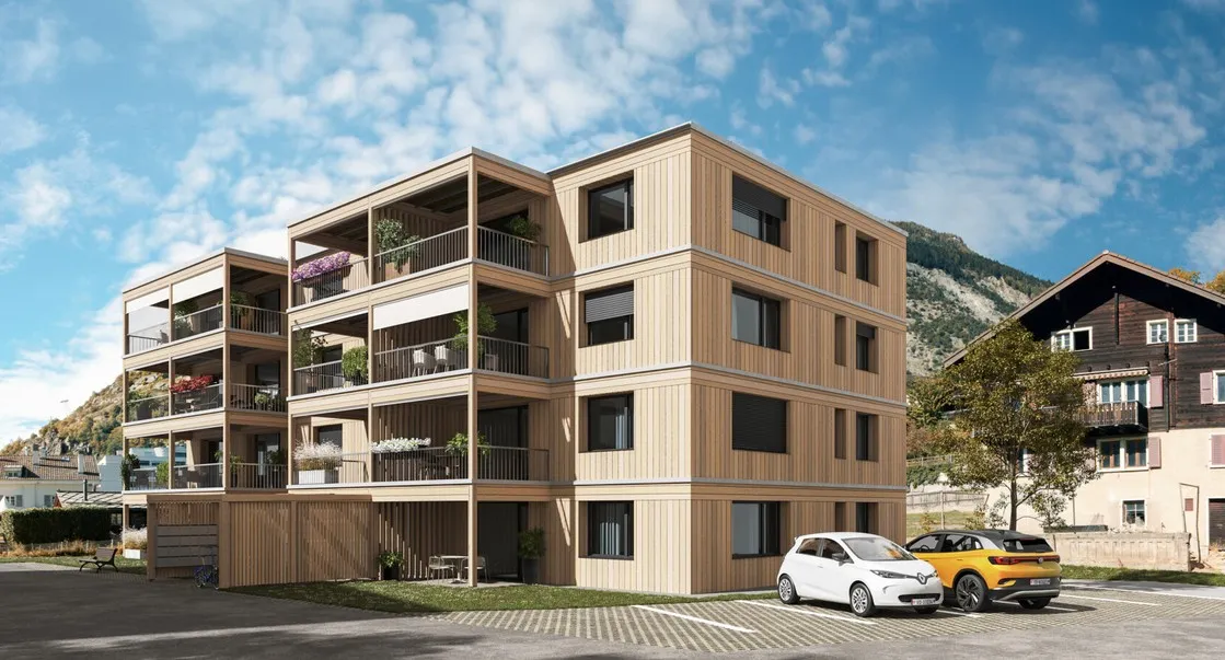 Visualisierung eines modernen Mehrfamilienhauses in Holzbauweise mit grossen Balkonen und begrüntem Dach mit Solaranlage. Das Gebäude fügt sich in eine alpine Dorflandschaft mit traditionellen Häusern, Strassen und einem bewaldeten Berghang im Hintergrund ein.