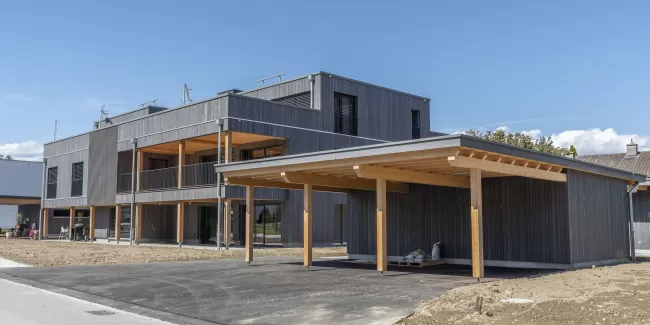 Mehrfamilienhaus Truberholzhaus Recherswil, Schweizer Holz, Truberholzwände sind nicht sichtbar, die Dübel sind nicht sichtbar, Fassade ist vorvergraut, es hat eine Attikawohnung, im Erdgeschoss und Obergeschoss hat es Loggaeinschnitte (Terrasse, Balkon), moderner Neubau, auf dem Dach ist ein Sonnensegel, Flachdach mit Solarpanel, es hat kein Keller dafür ein zusätzliches Gebäude mit Autounterstand und Lagerraum, Umschwung mit Rasen 