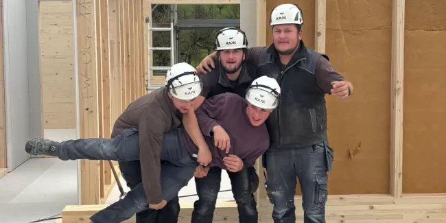 Vier Zimmerleute posieren lachend auf der Baustelle eines Holzhauses. Die fröhliche Stimmung zeigt den Teamgeist und die Freude am Handwerk bei der Zimmerei Hirschi AG.