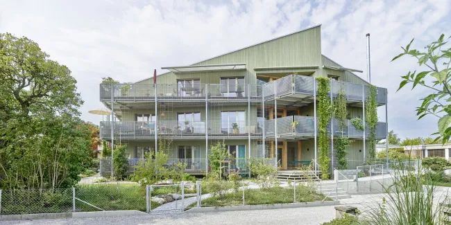 Aussenansicht eines modernen, mehrstöckigen Wohngebäudes in Holzbauweise mit hellgrüner Fassade. Das Haus verfügt über grosse Balkone mit Metallgeländern und Kletterpflanzen, die vertikal an den Fassadenelementen emporwachsen. Vor dem Gebäude liegt ein gepflegter Garten mit jungen Bäumen, Sträuchern und geschwungenen Wegen. Die Architektur wirkt offen und einladend, mit vielen Fenstern und einer klar strukturierten Formensprache. Im Vordergrund sind ein Kiesweg und ein Zaun sichtbar, während der Himmel leic