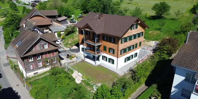 Luftaufnahme eines frisch renovierten, mehrstöckigen Holzhauses im traditionellen Stil mit brauner Schindelfassade und grünen Fensterläden. Das Gebäude steht inmitten einer ländlichen Umgebung mit Wiesen, Bäumen und benachbarten Wohn- und Wirtschaftsbauten. Vor dem Haus befindet sich ein neu angelegter Vorplatz, während rundherum noch Bereiche der Gartengestaltung und Bauarbeiten sichtbar sind. Im Hintergrund erstreckt sich eine grüne Hügellandschaft mit verstreuten Obstbäumen und einigen modernen Wohnhäuse