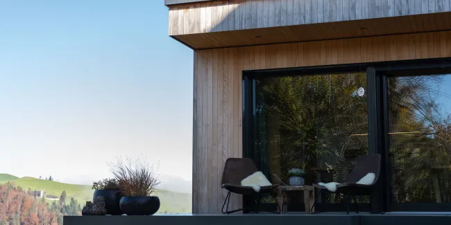 Aussenansicht eines modernen Holzhauses mit klarer Linienführung. Auf der erhöhten Terrasse stehen zwei Stühle mit Kissen und ein kleiner Tisch vor einer grossen Glasfront. Die Fassade besteht aus senkrechter Holzschalung, im Hintergrund sind Hügel, Bäume und eine ländliche Landschaft bei klarem Himmel zu sehen.