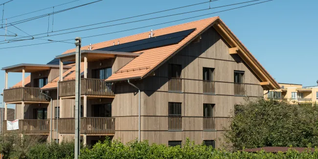 Aussenansicht eines mehrgeschossigen Wohnhauses in Holzbauweise mit Satteldach und integrierten Solarpanels. Die Fassade besteht aus senkrechter Holzschalung, die Balkone sind aus Holz gefertigt. Das Gebäude steht in einem Wohnquartier mit Hecken und Nachbarbauten, im Vordergrund verlaufen Bahnoberleitungen vor blauem Himmel.