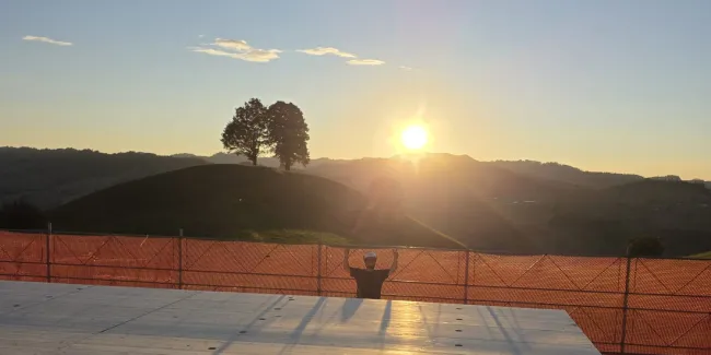 Blick über eine neu verlegte Holzdecke auf einer Baustelle bei Sonnenuntergang. Im Hintergrund steht ein Zimmermann am Gerüst vor der hügeligen Landschaft und hebt beide Arme in die Höhe, während die tief stehende Sonne über den Hügeln scheint.