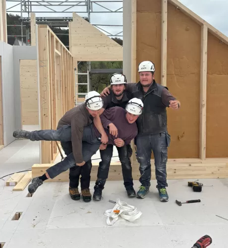 Vier Zimmerleute posieren lachend auf der Baustelle eines Holzhauses. Die fröhliche Stimmung zeigt den Teamgeist und die Freude am Handwerk bei der Zimmerei Hirschi AG.