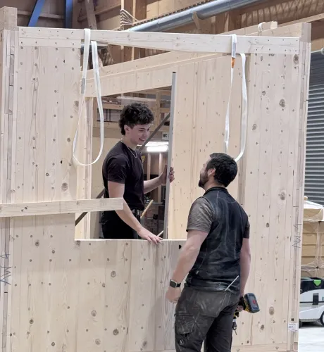 Zwei Mitarbeitende der Zimmerei Hirschi montieren in der Werkhalle ein vorgefertigtes Holzelement. Sie arbeiten an einer massiven Holzrahmenkonstruktion mit Fensteröffnung und stimmen sich dabei direkt vor Ort ab.