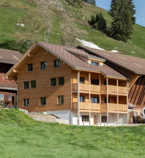 Aussenansicht eines neu errichteten Wohnhauses in Holzbauweise mit Satteldach und Balkonen, eingebettet in eine hügelige, ländliche Landschaft. Das Gebäude steht neben bestehenden landwirtschaftlichen Bauten. Im Vordergrund eine Wiese, im Hintergrund ein begrünter Hang mit Bäumen unter blauem Himmel.