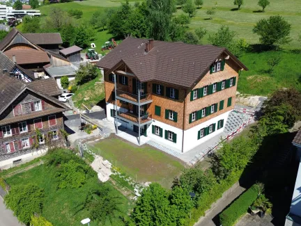 Luftaufnahme eines frisch renovierten, mehrstöckigen Holzhauses im traditionellen Stil mit brauner Schindelfassade und grünen Fensterläden. Das Gebäude steht inmitten einer ländlichen Umgebung mit Wiesen, Bäumen und benachbarten Wohn- und Wirtschaftsbauten. Vor dem Haus befindet sich ein neu angelegter Vorplatz, während rundherum noch Bereiche der Gartengestaltung und Bauarbeiten sichtbar sind. Im Hintergrund erstreckt sich eine grüne Hügellandschaft mit verstreuten Obstbäumen und einigen modernen Wohnhäuse