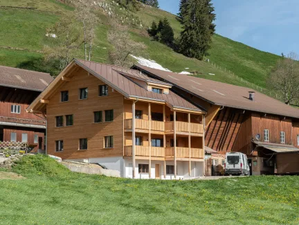 Aussenansicht eines neu errichteten Wohnhauses in Holzbauweise mit Satteldach und Balkonen, eingebettet in eine hügelige, ländliche Landschaft. Das Gebäude steht neben bestehenden landwirtschaftlichen Bauten. Im Vordergrund eine Wiese, im Hintergrund ein begrünter Hang mit Bäumen unter blauem Himmel.
