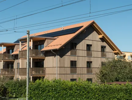 Aussenansicht eines mehrgeschossigen Wohnhauses in Holzbauweise mit Satteldach und integrierten Solarpanels. Die Fassade besteht aus senkrechter Holzschalung, die Balkone sind aus Holz gefertigt. Das Gebäude steht in einem Wohnquartier mit Hecken und Nachbarbauten, im Vordergrund verlaufen Bahnoberleitungen vor blauem Himmel.